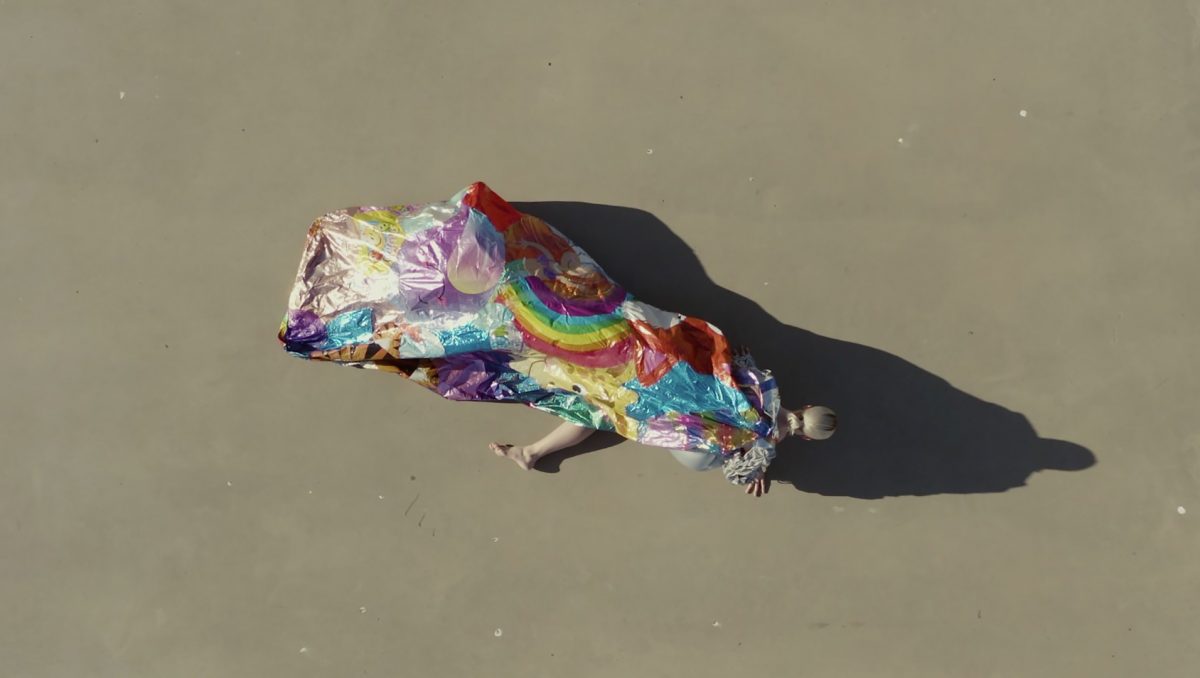 In this aerial perspective by Zoe Bastin and part of the Conflated exhibition the artist is stretched across an outdoor concrete ground and mostly obscured by a mass of brightly coloured balloons melded together as one inflatable object.