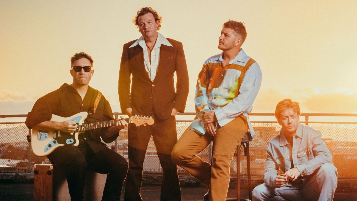 Four band members in row, one holds a guitar, on a hill with sunset behind.