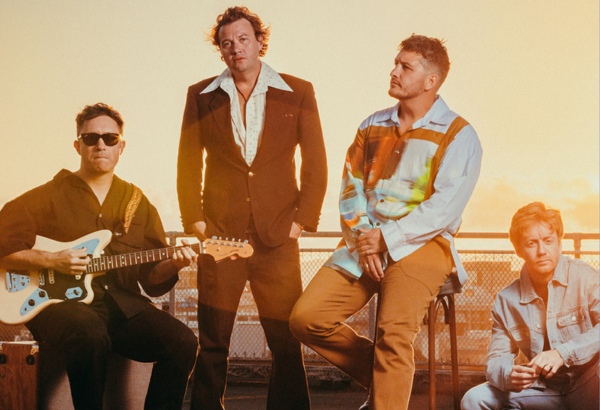 4 band members in a row, one holds a guitar, sunset on hills behind them.